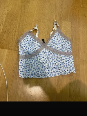 Blommig spetslinnetopp från Divided - Supersöt blommig linnetopp från Divided med blått och vitt mönster. Toppen har spetsdetaljer i grått och smala axelband. Perfekt för sommarens alla outfits och ger en romantisk vibe.
