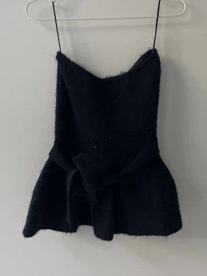 Svart stickad axelbandslös klänning Zara - Supersnygg svart stickad klänning från Zara med axelbandslös design och knyt i midjan. Kort modell som ger en trendig siluett och passar perfekt till lager på lager. Mjuk och lite fluffig känsla i materialet.
