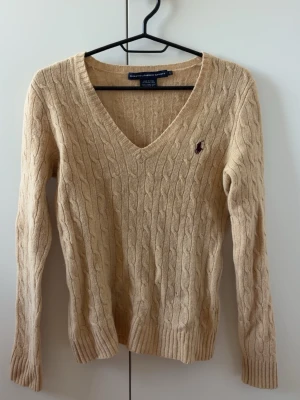 Beige kabelstickad tröja Polo Ralph Lauren - Kabelstickad tröja i beige från Polo Ralph Lauren med V-ringning och lång ärm. Tröjan har det klassiska broderade logotypen på bröstet och ribbade muddar vid ärmslut och nederkant. Perfekt för lager på lager och enkel att styla.