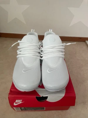 Nike vita sneakers med mesh - Fräscha helvita Nike sneakers med mesh-ovandel och klassisk swoosh på sidan. Skorna har rund tå, vita snören och en bekväm, vadderad sula. Perfekta för dig som gillar stilrena och sportiga sneakers.