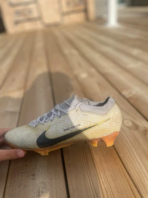 Nike vita fotbollsskor med gulddetaljer - Snygga vita fotbollsskor från Nike med gulddetaljer och svart swoosh. Skon har snörning, låg profil och en yttersula med orange och gröna inslag samt Zoom-teknologi för extra respons. Perfekt grepp för spel på gräsplan.