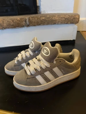Grå Adidas sneakers - Snygga grå sneakers från Adidas med klassiska vita ränder på sidorna och Adidas-logga på plösen och hälen. Skorna är låga och har breda vita skosnören. Ovandelen är i mjuk mocka och sulan är vit med beige inslag.