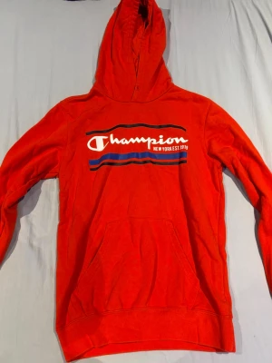 Röd Champion hoodie med tryck - Snygg röd hoodie från Champion med stort vitt och blått tryck på bröstet där det står 'Champion New York EST 1919'. Hoodien har huva och en stor magficka framtill. Perfekt för en avslappnad streetwear-look.
