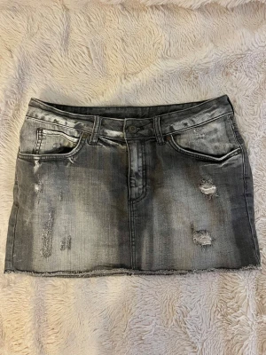 Grå jeanskjol med slitningar - Säljer en grå kort jeanskjol med fina detaljer som trendar högt just nu. Har inga defekter och är perfekt nu i sommar.