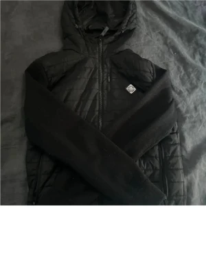 J Lindberg cardigan - Snygg svart cardiganjacka med quiltad front och stickade ärmar. Jackan har dragkedja, huva och en liten logotyp på bröstet. Perfekt för lager-på-lager den är är fin skick storlek S om intresse skriv elle mer bilder
