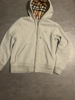 Grå Burberry hoodie m - Snygg grå zip-hoodie med klassiskt rutigt foder i huvan. Hoodien har dragsko, två fickor framtill och ribbade muddar vid ärmar och nederkant. Perfekt för en chill och stilren look. Materialet ser ut att vara mjuk bomullsblandning.