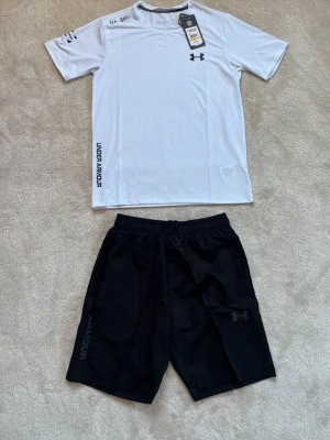 Under Armour träningsset svart/vit - Snyggt träningsset från Under Armour med svarta shorts och vit t-shirt. Shortsen har elastisk midja och logga på benet, t-shirten har tryck på bröstet och sidan. Båda i lätt och snabbtorkande material, perfekt för träning.