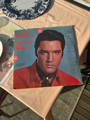 Elvis Presley LP A Portrait in Music - Vinylskiva med Elvis Presley, 'A Portrait in Music'. Omslaget har ett porträtt av Elvis i röd skjorta och svart scarf. Inuti finns gul text med information om albumet och låtarna. Perfekt för samlare eller Elvis-fans.