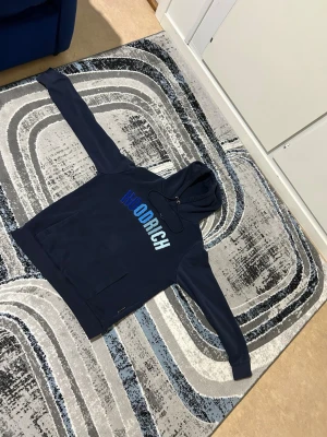 Mörkblå HOODRICH hoodie med tryck - Snygg mörkblå hoodie från HOODRICH med stor logga i blå toner på bröstet. Klassisk huva med dragsko och stor magficka framtill. Perfekt för en chill och avslappnad stil. Mjuk och bekväm bomullsblandning.