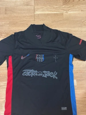 Svart FC Barcelona x Nike tröja - Säljer en svart FC Barcelona tröja från Nike med Cactus Jack-grafik på bröstet. Tröjan har klubbmärke i svart, röda och blå detaljer på sidorna samt tryck på ärmen. Perfekt för dig som gillar unika fotbollströjor och streetwear.