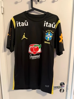 Svart Brasilien tränings t-shirt - Snygg svart tränings t-shirt från Brasiliens landslag med gula detaljer och flera tryckta loggor, bland annat CBF-emblemet, Jordan-loggan och sponsorer som Guaraná Antarctica och Itaú. Tillverkad i ett lätt och ventilerande material, perfekt för fotbollsträning.