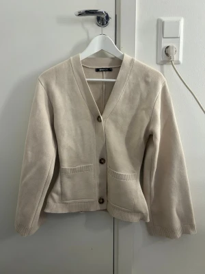 Beige cardigan från Gina Tricot - Stilren beige kofta från Gina Tricot med v-ringning och två stora knappar framtill. Koftan har två fickor fram och ribbade muddar vid ärmslut och nederkant. Perfekt att slänga över en t-shirt för en chill look.
