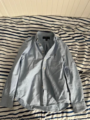 Ljusblå skjorta från Ralph Lauren - Klassisk ljusblå skjorta från Ralph Lauren med knappar framtill och lång ärm. Skjortan har en liten broderad logga på bröstet och är tillverkad i mjuk bomull. Perfekt för en stilren och avslappnad look.Kan gå ner vid snabb affär