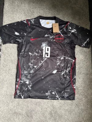 Svart Kanada matchtröja Nike - Säljer en svart Kanada matchtröja från Nike med vitt och rött stänkmönster, nummer 19 och namnet Davies på ryggen. Tröjan har korta ärmar, röd Nike-logga och Kanada-emblem på bröstet. Perfekt för dig som älskar fotboll och vill sticka ut på planen.