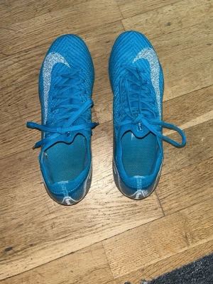 Blå Nike fotbollsskor med dobbar - Säljer ett par blå Nike fotbollsskor med vita detaljer och dobbar för spel på gräs. Skorna har snörning och är tillverkade i ett lätt syntetmaterial som ger bra bollkontroll och passform. Perfekta för dig som vill ha stilrena och funktionella skor på planen.