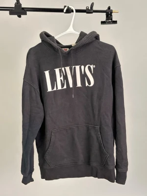 Svart hoodie från Levi's med tryck - Svart hoodie från Levi's med stort vitt logotryck på bröstet. Modellen har huva med dragsko och en stor magficka framtill. Avslappnad passform och mjukt material som känns skönt mot huden.