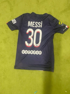 Mörkblå PSG Messi matchtröja - Säljer en mörkblå PSG matchtröja med Messi och nummer 30 på ryggen. Tröjan har korta ärmar, vita tryck och detaljer i rött och vitt. Perfekt för dig som älskar fotboll och vill stötta ditt favoritlag på planen eller läktaren.