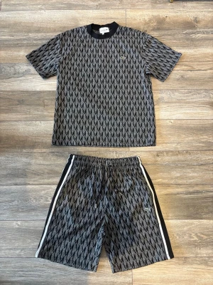Svart och grått Lacoste sett  - Snyggt tvådelat Lacoste set med t-shirt och shorts i svart och grått med diamantmönster. Perfekt för en avslappnad och trendig look.