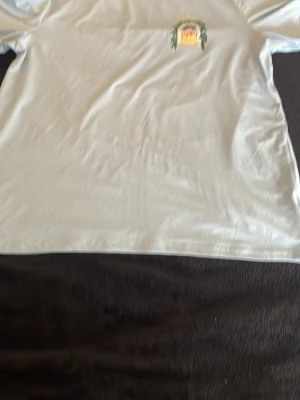 Vit t-shirt med tryck Casablanca - Säljer en vit t-shirt med färgglatt tryck på ryggen som föreställer en byggnad och texten 'Avenida Casablanca'. T-shirten har korta ärmar och en klassisk passform. Perfekt för dig som gillar grafiska motiv och vill sticka ut lite extra.
