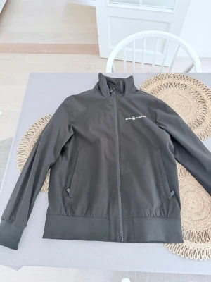 Svart vindjacka från Sail Racing - Stilren svart vindjacka från Sail Racing med dragkedja framtill och två snedställda fickor med knappstängning. Jackan har hög krage och diskret logga på bröstet. Perfekt för blåsiga dagar och enkel att matcha med olika outfits. Den ser rödaktig ut på grund av kameran men är helt svart utan några skador väldigt bra skick