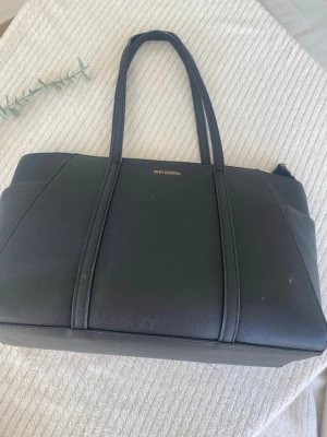 Svart handväska från Don Donna - Stilren svart handväska från Don Donna i slätt konstläder. Väskan har två långa handtag och en rymlig, rektangulär form. Perfekt för dig som vill ha en klassisk och enkel look med plats för allt du behöver.