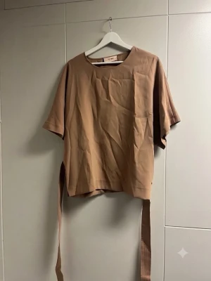 Beige blus från Mos Mosh - Snygg beige blus från Mos Mosh med rund halsringning och korta ärmar. Tillverkad i ett mjukt, lätt material och har ett matchande midjeband som kan knytas för en mer markerad siluett. Perfekt för en stilren och avslappnad look.