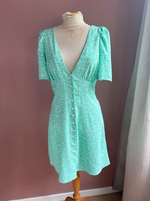 Mintgrön kortklänning med knappar - Superfin mintgrön klänning i stl S med vitt blommönster, v-ringning och korta puffärmar. Klänningen har knappar framtill och en figurnära passform som ger en feminin siluett. Perfekt för vår och sommar!