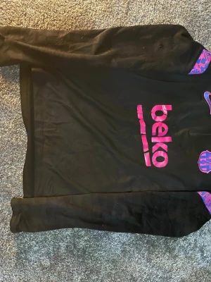 Svart Nike träningströja FC Barcelona - Svart långärmad träningströja från Nike med FC Barcelona-logga och rosa BEKO-tryck på bröstet. Tröjan har Dri-FIT-material som andas och detaljer i lila och rosa på ärmarna. Perfekt för fotbollsträning eller gym.