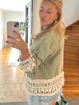 Grön kort jacka med virkade detaljer - Säljer en ljusgrön, kort jacka med stora knappar framtill och unika virkade detaljer med tofsar längs ärmslut och nederkant. Jackan har en avslappnad passform och är perfekt för dig som vill sticka ut med din stil. Materialet ser ut att vara bomull eller denim.