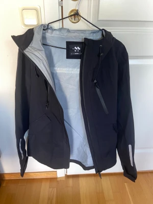 Shell jacket "Ws Everyday Jacket", Black, M Lager 157 - Shell jacket "Ws Everyday Jacket", Black, M med grått innerfoder och huva. Jackan har dragkedja framtill, två sidofickor med dragkedja och justerbar huva med snörning. Reflekterande detaljer på ärmarna och sportig design. Perfekt för blåsiga dagar. 