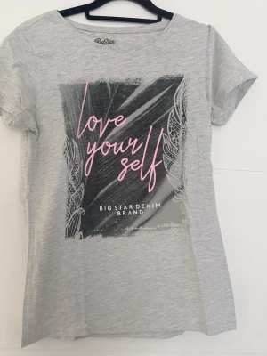 Grå t-shirt med tryck från Big Star - Snygg grå t-shirt från Big Star med ett stort grafiskt tryck framtill och texten 'love yourself' i rosa. T-shirten har rund halsringning och korta ärmar. Perfekt för en avslappnad och trendig look.