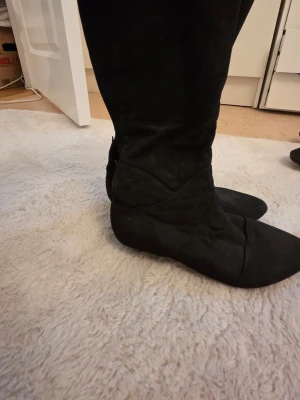 Svarta knähöga stövlar i mocka-look - Snygga svarta stövlar med spetsig tå och knähögt skaft. Tillverkade i ett material som liknar mocka och har en stilren, slimmad design utan synliga detaljer. Perfekta för att lyfta din outfit med en trendig touch.