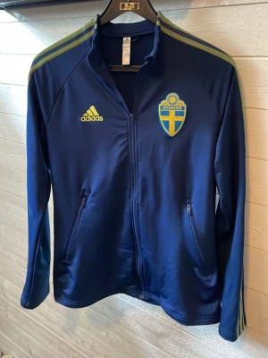 Sverige träningsjacka - Mörkblå träningsjacka från Adidas med Sveriges fotbollslandslagsmärke på bröstet. Jackan har hel dragkedja, två fickor med dragkedja och gula detaljer på axlarna samt Adidas-logga. Perfekt för fotbollsträning eller supporterliv.