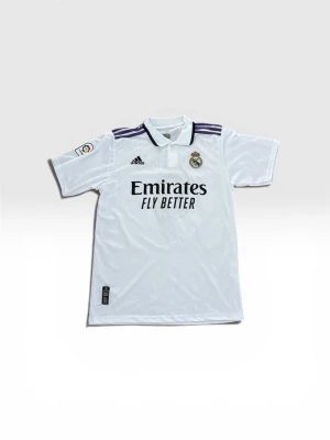 Real Madrid matchtröja Benzema 9 - Snygg vit Real Madrid matchtröja från Adidas med Benzema och nummer 9 på ryggen. Tröjan har klubbmärke, sponsortryck och La Liga-märke på ärmen. Klassisk pikékrage och lila detaljer på axlarna. Perfekt för fotbollsfans!