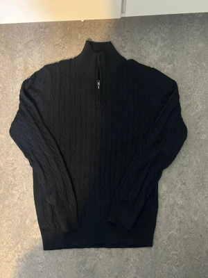 Mörkblå kabelstickad tröja Dressmann - Mörkblå kabelstickad tröja från Dressmann med halv dragkedja och hög krage. Tröjan har långa ärmar och ribbade muddar vid ärmslut och nederkant. Perfekt för dig som gillar klassisk och stilren look.