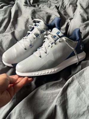 Gråa sneakers från Under Armour - Snygga sneakers från Under Armour i ljusgrått med mörkblå detaljer och vit sula. Skorna har en sportig design med mönstrad yttersula för extra grepp och perforerade partier för ventilation. Perfekta för träning eller vardagsaktiviteter.
