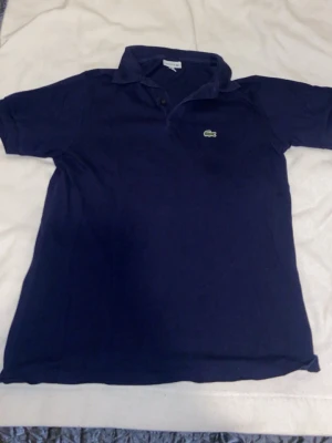 Marinblå pikétröja från Lacoste - Snygg marinblå pikétröja från Lacoste med klassisk krage och knappar framtill. Tröjan har det ikoniska krokodilmärket broderat på bröstet och är tillverkad i ett mjukt bomullsmaterial. Perfekt för en stilren och sportig look.