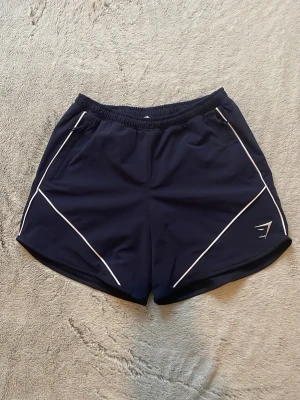 RETRO SHORTS från gymshark! - RETRO SHORTS, Regular Fit! Mörkblå sportshorts med vita linjer längs sidorna och elastisk midja. Lätta och bekväma, perfekta för träning eller annat! Skriv vid frågor eller pris! 