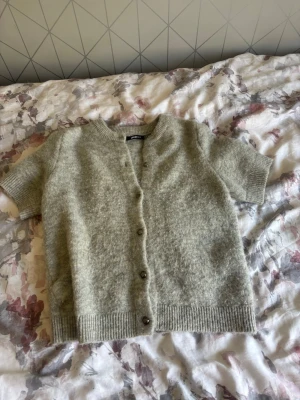 Ljusgrå kortärmad kofta - Kortärmad kofta i ljusgrått, stickad i mjukt material med rund halsringning och ribbade kanter. Knäpps framtill med fyra knappar. Perfekt för lager på lager och en chill look. Lite nopprig 
