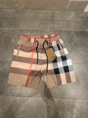 Rutiga badbyxor från Burberry - Säljer ett par snygga badbyxor från Burberry med klassiskt rutigt mönster i beige, brunt, svart, vitt och rött. De har resårmidja med dragsko och sidofickor. Perfekta för stranden eller poolen och tillverkade i snabbtorkande material.