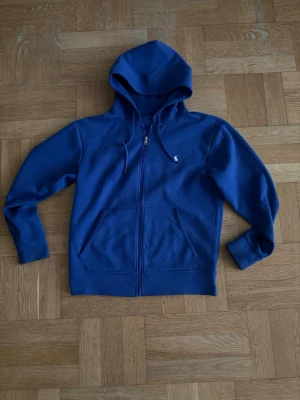 Blå zip hoodie från Polo Ralph Lauren - Snygg blå hoodie med dragkedja från Polo Ralph Lauren. Klassisk design med huva och dragsnören, två fickor framtill och den ikoniska vita loggan broderad på bröstet. Tillverkad i mjuk bomullsblandning som är skön att ha på sig.