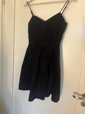 Svart kort klänning med smala band - En svart kort klänning med smala axelband och v-ringad urringning. Klänningen har en enkel, stilren design och är lätt utställd nedtill. Perfekt för dig som gillar minimalistisk stil och vill ha något enkelt men snyggt.
