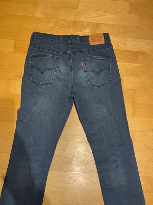 Mörkblå raka jeans från Levi's - Säljer ett par klassiska mörkblå jeans från Levi's med raka ben och femficksmodell. Jeansen har tydliga sömmar, läderpatch bak i midjan och Levi's röda etikett på bakfickan. Tillverkade i slitstarkt denimtyg, perfekta till alla tillfällen.