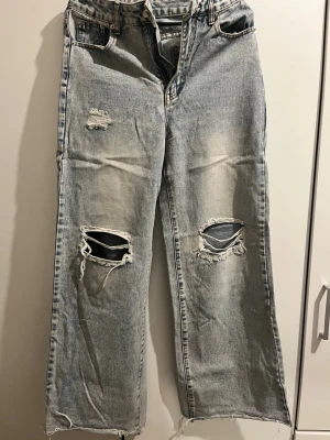 Ljusa slitna raka jeans - Säljer ett par ljusblå jeans med raka ben och flera slitningar och hål på framsidan. Jeansen har hög midja och råa kanter vid bensluten för en trendig look. Perfekta för dig som gillar en avslappnad och edgy stil.