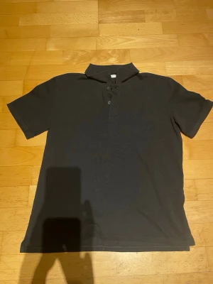 Svart pikétröja med krage - Klassisk svart pikétröja med korta ärmar och krage. Tröjan har knappar framtill och är tillverkad i ett mjukt material som passar perfekt till många olika stilar. Enkel och stilren design som funkar till det mesta.