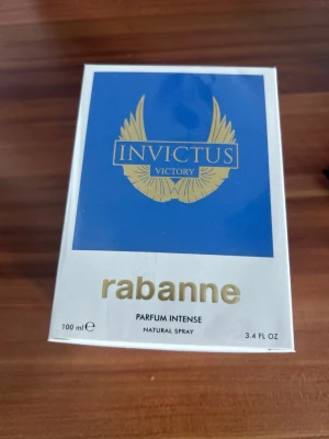 Invictus Victory Parfum Intense 100 ml - Parfymen Invictus Victory Parfum Intense från Rabanne kommer i en elegant vit och blå förpackning med guldiga detaljer. Flaskan rymmer 100 ml och är en naturlig spray. Designen är modern och lyxig med tydlig logotyp och vingar i guld.