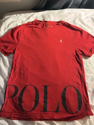 Röd t-shirt med stor POLO-logga - Snygg röd t-shirt med stor svart POLO-logga längst ner och liten broderad logga på bröstet. Klassisk rund hals och korta ärmar. Tillverkad i mjuk bomull som känns skön mot huden. Perfekt för dig som gillar stilrena plagg med tydlig branding.