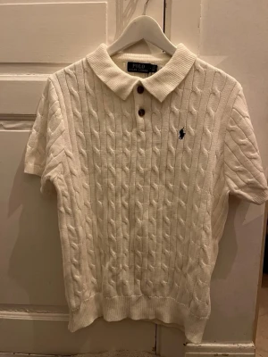 Vit kabelstickad pikétröja Polo Ralph Lauren - Vit kabelstickad pikétröja från Polo Ralph Lauren med korta ärmar och klassisk krage. Tröjan har två bruna knappar framtill och den ikoniska loggan broderad på bröstet. Perfekt för en preppy look.Priset går att diskutera vid snabb affär, finn de frågor så är de bara o skriv 😊