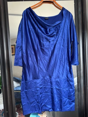 Kornblå kort klänning med drapering - Snygg kornblå klänning i kort modell med draperad halsringning och trekvartsärm. Tillverkad i ett glansigt material som ger en elegant look. Perfekt för dig som vill sticka ut med färg och stil.