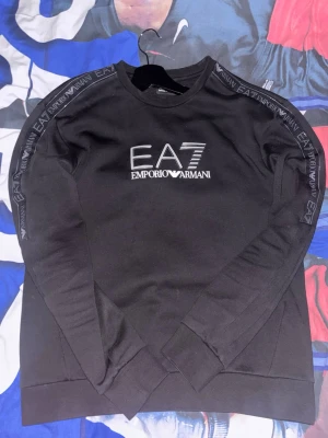 Svart EA7 Emporio Armani sweatshirt - Svart sweatshirt från EA7 Emporio Armani med stort logotryck på bröstet och logoband längs ärmarna. Tröjan har rund halsringning och långa ärmar. Perfekt för en stilren och sportig look. Storleken är i L men passar mer M.Priset går alltid att diskutera!